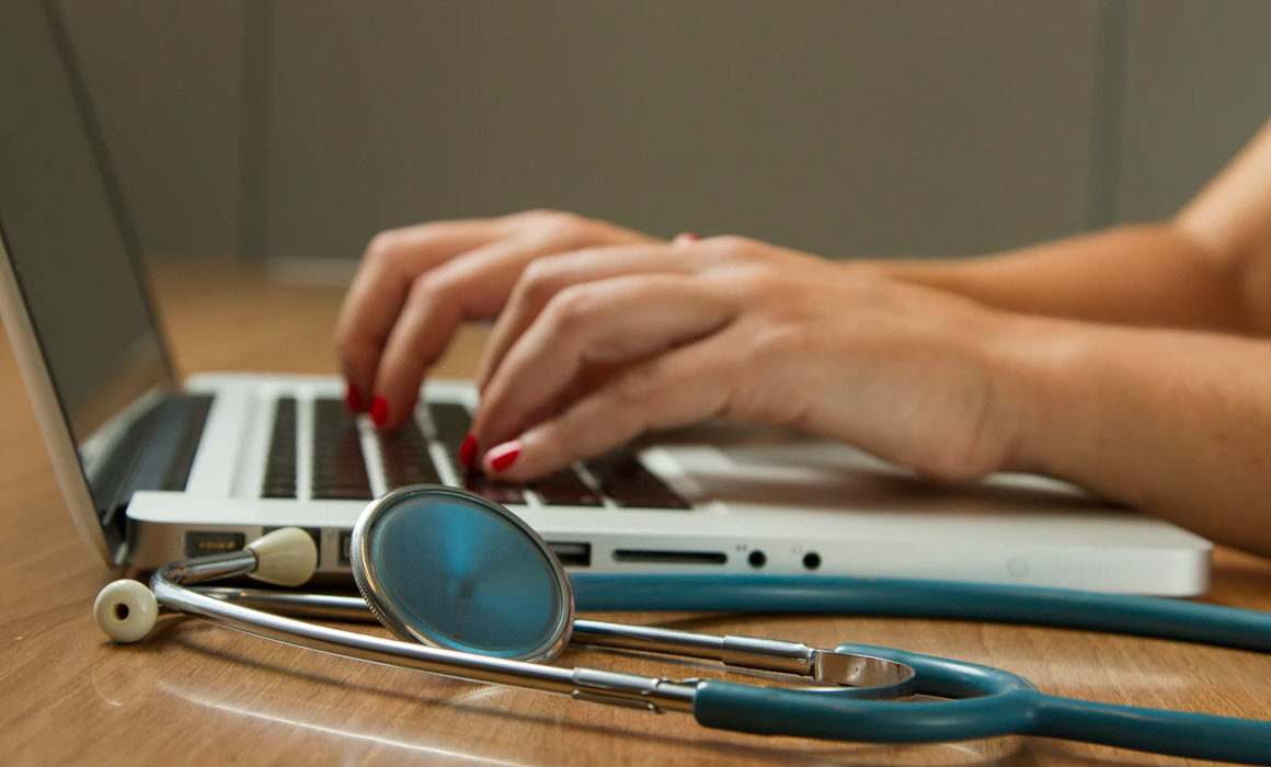 Hands typing on a laptop with a stethoscope in view.