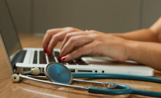 Hands typing on a laptop with a stethoscope in view.