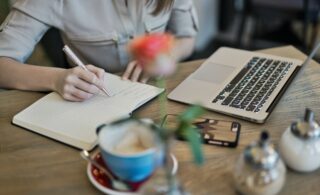 Woman sat at a table, writing in a notebook, coffee cup, laptop, salt & pepper shaker and a vase with a single orange rose slightly out of focus