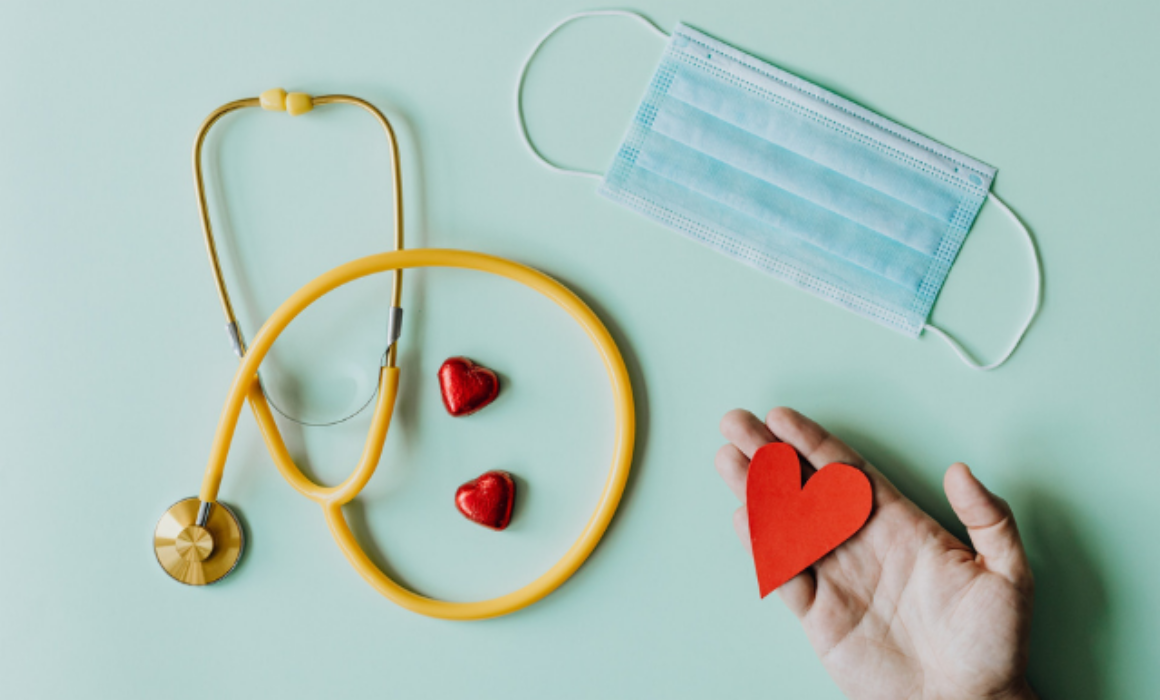 Stethascope, face mask and a hand holding a little red paper heart on a pale blue background