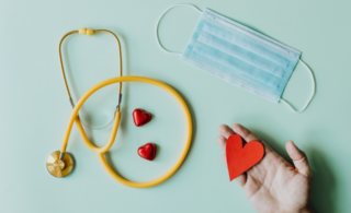 Stethascope, face mask and a hand holding a little red paper heart on a pale blue background