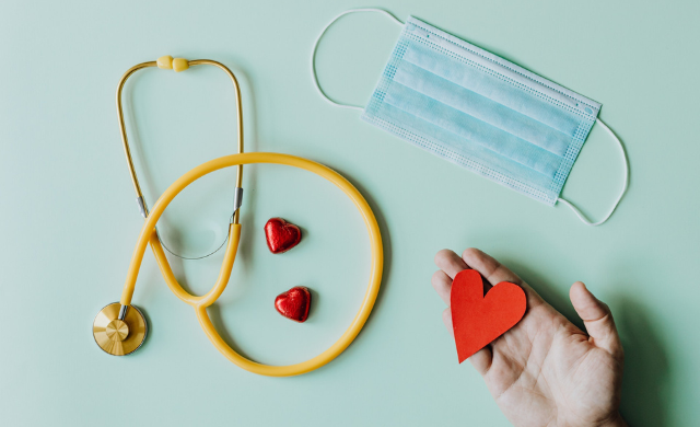 Stethascope, face mask and a hand holding a little red paper heart on a pale blue background